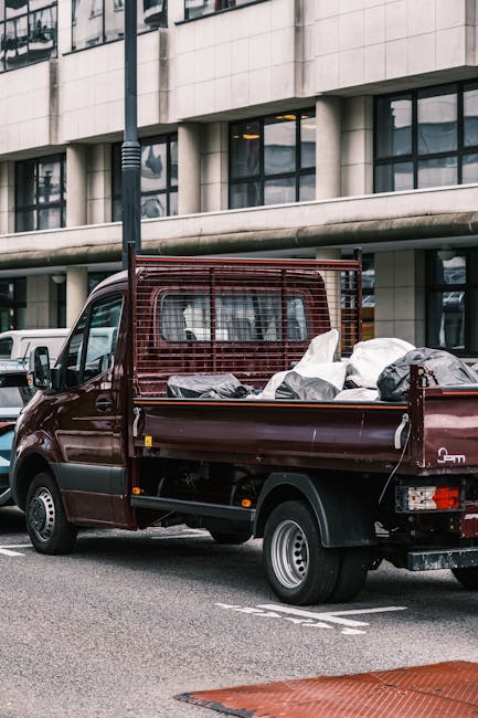 A burgundy flatbed pickup truck parked on an urban street in front of a modern glass-fronted building with large windows and a curved facade. The truck's bed is filled with miscellaneous rubbish, including several large white and black plastic bags and loose debris. The vehicle features a protective metal grille at the cab's rear, and its surface looks clean but used. The scene is illuminated by natural daylight, with a black streetlamp positioned behind the truck. Surrounding the truck, there are other parked vehicles, and the street markings suggest a designated parking area. The setting appears to be a city environment where independent waste collection services, such as those by Shepherds Bush rubbish removal, operate to facilitate private disposal of rubbish outside of municipal collection systems.