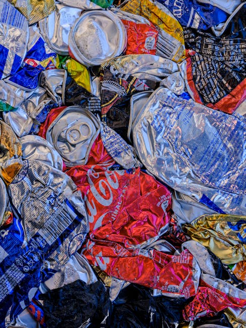 A large pile of crushed and crumpled aluminum cans, primarily made of soft drink and beverage containers, with visible ridges, pull-tabs, and logos such as Coca-Cola. The cans display metallic silver surfaces with areas of red, blue, yellow, and black printing, some partially flattened or torn, scattered chaotically in a mixed arrangement. The cans' textures vary from smooth and shiny to dented and rough due to crushing, and they are piled on a surface that appears to be outdoors or in a waste disposal area. Surrounding the cans, there are hints of other waste materials, but the cans dominate the visual scene, indicating a collection or disposal of recyclable aluminum materials. The scene exemplifies typical waste handling associated with independent rubbish clearance services like those conducted by Shepherds Bush waste management specialists, emphasizing the importance of proper rubbish removal and recycling. The lighting highlights the reflective surfaces of the aluminum and emphasizes the worn, crumpled state of the cans, situating the image within the context of private waste collection or on-site clearance processes.