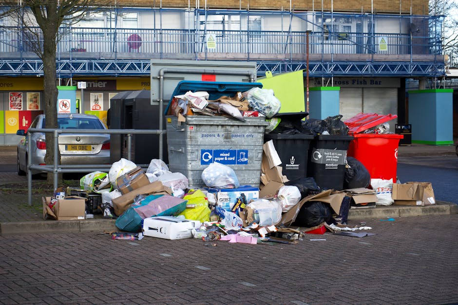A large pile of mixed household waste and rubbish is overflowing from several black and grey garbage bins, along with a red recycling bin, on a paved urban street near parked cars. The waste includes various materials such as cardboard boxes, plastic bags, paper, and packaging debris, some of which are scattered on the ground in front of the bins. The grey bin, branded with the text 'mixed paper & card,' is partially open, revealing crumpled papers, cardboard, and miscellaneous refuse inside, while the black bins contain bags of rubbish and other waste items. In the background, a building with blue scaffolding and a row of commercial storefronts is visible, indicating a retail or public area. The scene reflects a typical instance of improperly disposed waste, which can be relevant to private rubbish removal services like those offered by wasteclearanceshepherdsbush.co.uk for on-site clearance or alternative waste handling outside standard council rubbish collections.