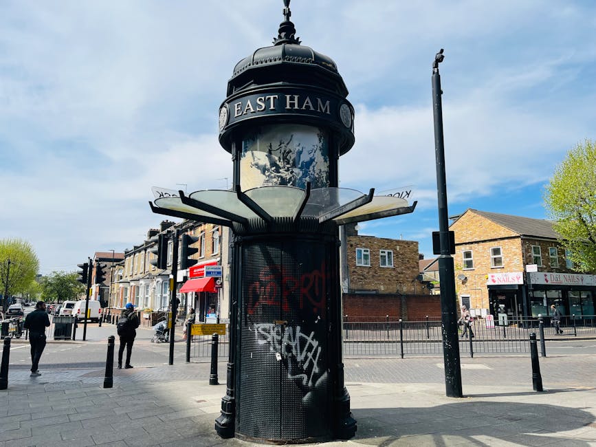 A cylindrical information kiosk with a domed top, painted black, situated on a busy city street corner during daytime. The kiosk features a glass display case at the top with the text 'EAST HAM' visible on a dark band encircling the structure. Surrounding the base of the kiosk is a metal grate with graffiti tags in white and red spray paint. To the left and right, black metal poles with small barriers are installed along the pavement, guiding pedestrian movement. In the background, several people are walking, some with backpacks, and there are brick and stone buildings, shops including a nail salon and a dry cleaners, and traffic lights at the intersection. The sky above is partly cloudy with patches of blue, and the trees lining the street have fresh green leaves, suggesting springtime. The scene depicts a typical urban environment where independent rubbish or waste clearance services could operate nearby, with the street infrastructure reflecting a well-maintained public space in Shepherds Bush, London.