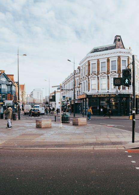 A busy urban street corner featuring a prominent three-storey corner building with a curved façade, white decorative architectural details, and large windows, housing a pub named 'The World's End' on the ground floor. The street scene includes pedestrians walking on the sidewalk, some crossing the intersection, and vehicles such as cars and a bus moving along the road. The foreground shows a wide, paved pedestrian crossing with wooden planters positioned at the intersection edges, and traffic lights with green pedestrian signals. The sky is partly cloudy, and typical street lighting fixtures are visible above the road. The scene captures the lively atmosphere of city life, with a mix of commercial and pedestrian activity, reflecting urban waste management context related to on-site clearance or private rubbish collection in busy high street environments, as managed by waste clearance services like those offered by wasteclearanceshepherdsbush.co.uk.